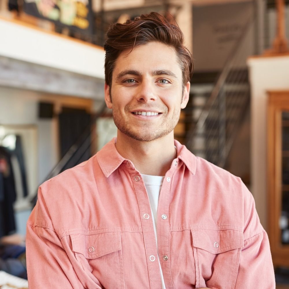 Portrait Of Smiling Male Owner Of Fashion Store Standing In Front Of Clothing Display